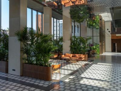 Słoneczny korytarz hotelowy z zielonymi roślinami i drewnianymi ławkami na wzorzystej podłodze.