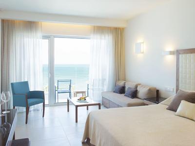 Chambre d'hôtel lumineuse avec lit double, canapé, chaise bleue et balcon avec vue sur la mer.