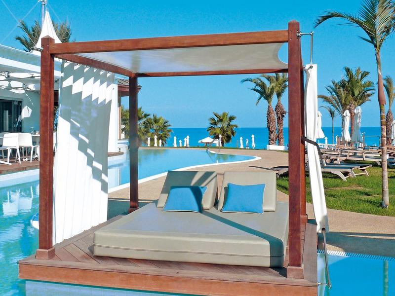 Lit d'extérieur moderne avec cadre en bois donnant sur la piscine et la mer sous un ciel bleu.