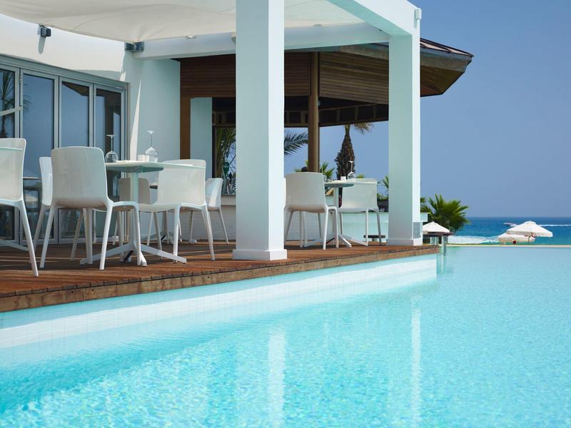 Espace piscine moderne avec chaises et tables blanches sur sol en bois sous un auvent.