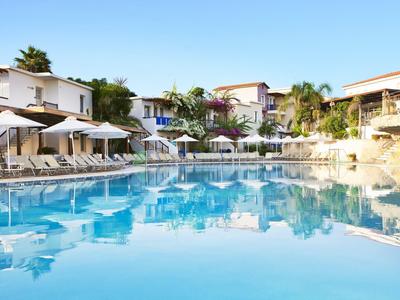 Großer blauer Pool mit Liegestühlen und Strandschirmen, umgeben von weißen Gebäuden und Palmen.
