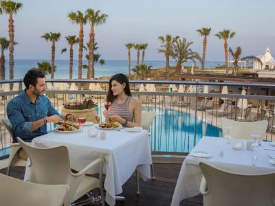 Paar isst draußen am gedeckten Tisch mit Blick auf Pool, Palmen und Meer bei klarem Himmel.