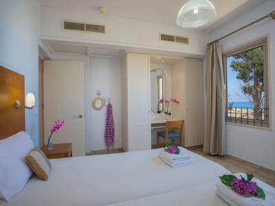 Helles Schlafzimmer mit großem Bett, Fenster mit Blick auf Palmen, Holzmöbeln und weißen Wänden.