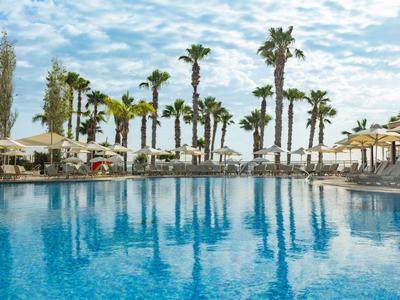 Großer Pool mit klar blauem Wasser, umgeben von Palmen und Sonnenschirmen unter blauem Himmel.