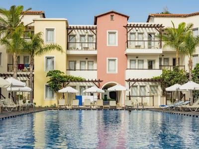Hotel con piscina al aire libre, tumbonas y sombrillas, rodeado de palmeras y cielo despejado.