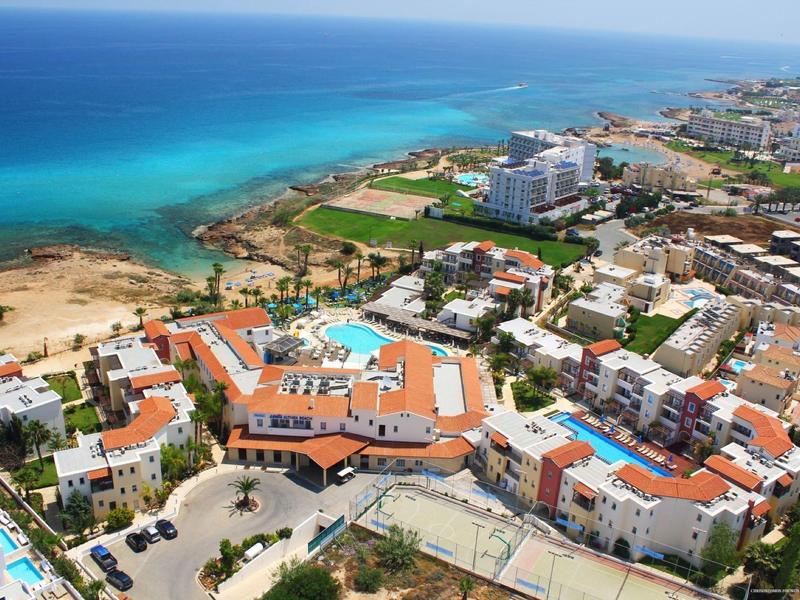Küstenstadt mit orangefarbenen Dächern, blauen Pools und türkisfarbenem Meer im Hintergrund.