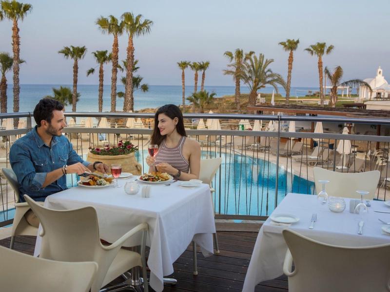 Paar isst draußen am gedeckten Tisch mit Blick auf Pool, Palmen und Meer bei klarem Himmel.