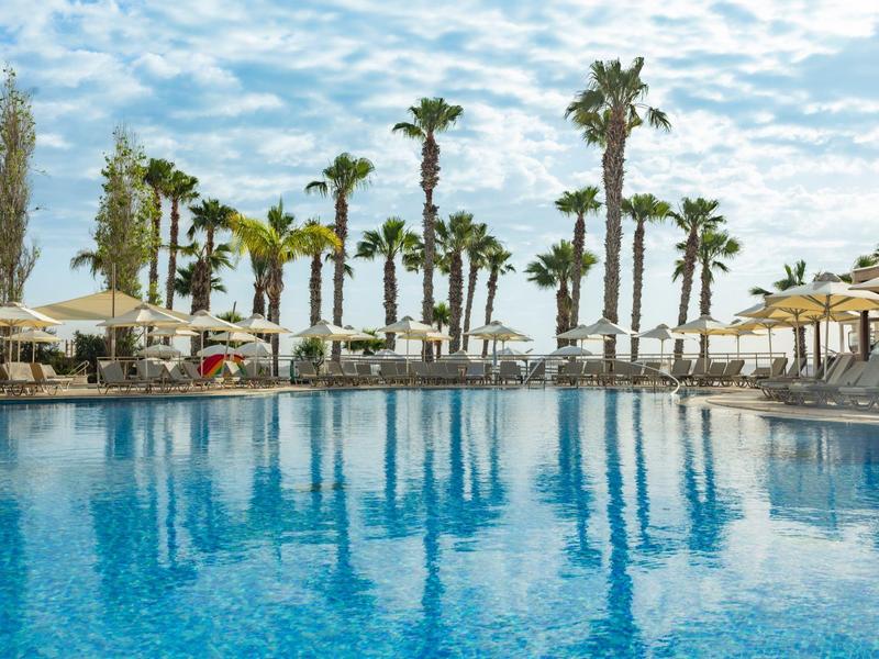 Großer Pool mit klar blauem Wasser, umgeben von Palmen und Sonnenschirmen unter blauem Himmel.