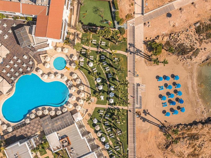Luftaufnahme eines Resorts mit Pool, Sonnenschirmen, Grünflächen und angrenzendem Sandstrand am Meer.