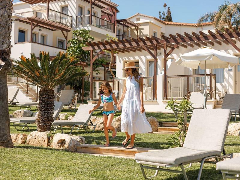 Zwei Frauen im Sommerkleid gehen über grünen Rasen vor einem weißen Haus mit Pergola.