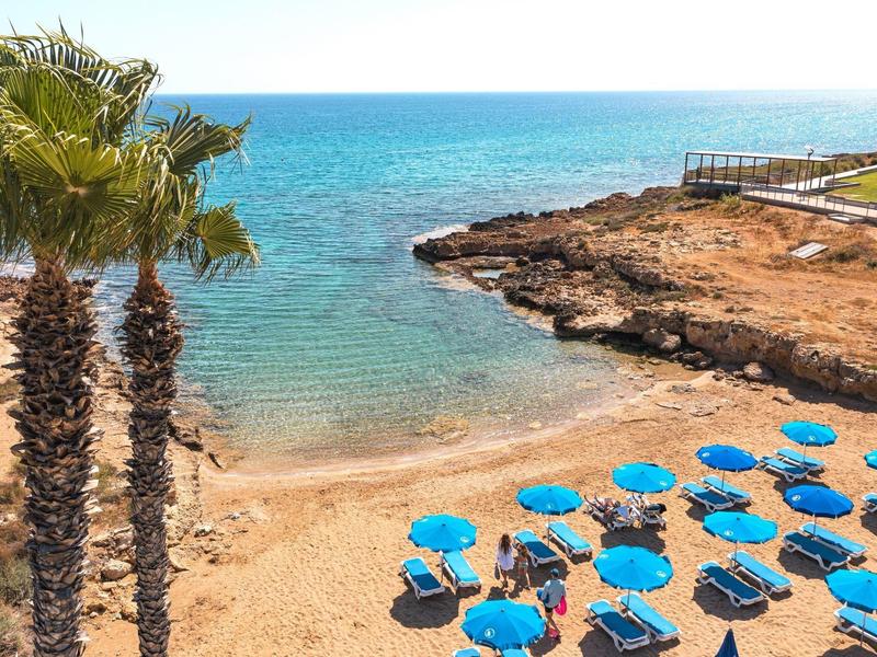 Strand mit blauem Sonnenschirmen, Palmen und felsiger Küste am klaren, blauen Meer unter hellem Himmel.
