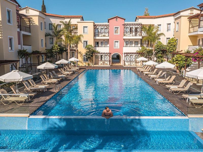 Großer blauer Pool mit weißen Liegestühlen und Sonnenschirmen vor hellen mediterranen Gebäuden.