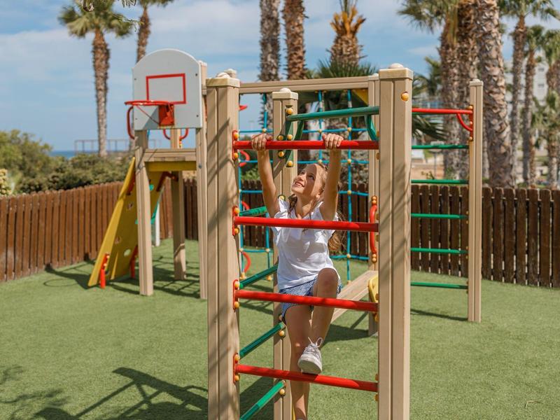 Kind spielt auf bunter Klettergerüst mit Basketballkorb und Palmen im Hintergrund.