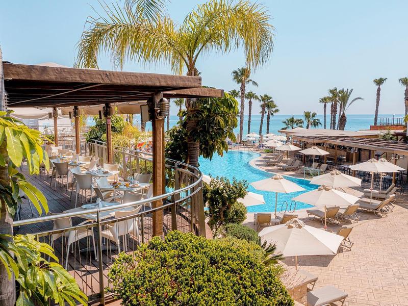 Strandhotel mit Pool, weißen Sonnenschirmen, Palmen und einer Terrasse mit Sitzplätzen und Glasgeländer.