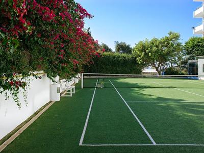 Kleiner Tennisplatz mit grünem Kunstrasen, umgeben von Blumen und Hotelgebäuden.