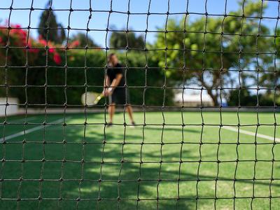 Tennisplatz mit grünem Boden, Netz im Vordergrund und Spieler bei sonnigem Wetter.