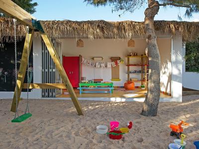 Kinderspielplatz mit Sand, Schaukel, Sandspielzeug und bunten Möbeln vor einem weißen Gebäude.