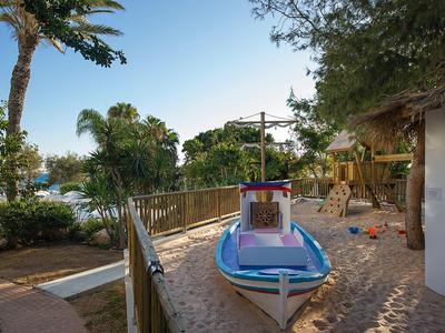Kleiner Kinderboot-Spielplatz am Strand neben einem Hotel, umgeben von Palmen und Pflanzen.