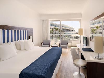 Helles Hotelzimmer mit großem Bett, blauen Akzenten und Balkon mit Sitzgelegenheiten.