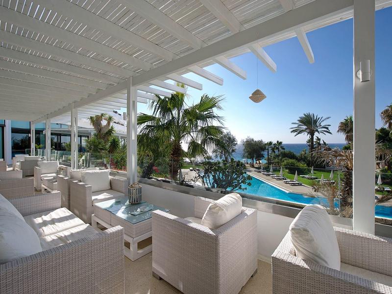 Überdachte Terrasse mit weißen Möbeln und Blick auf Pool und Palmen unter blauem Himmel.