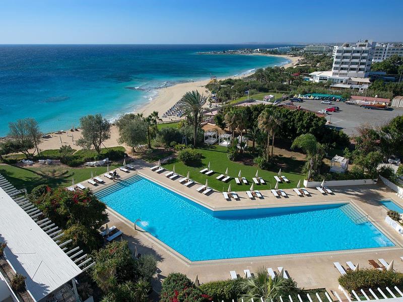 Blick auf einen Hotelpool mit Liegestühlen, Palmen und einem Sandstrand am klaren blauen Meer.