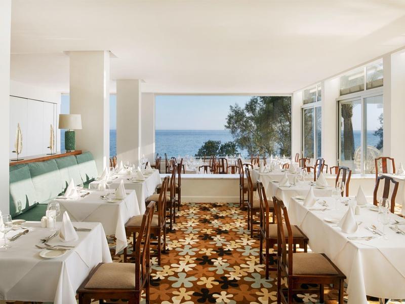 Eleganter Speisesaal mit weißen Tischen, Stühlen und Blick aufs Meer durch große Fenster.
