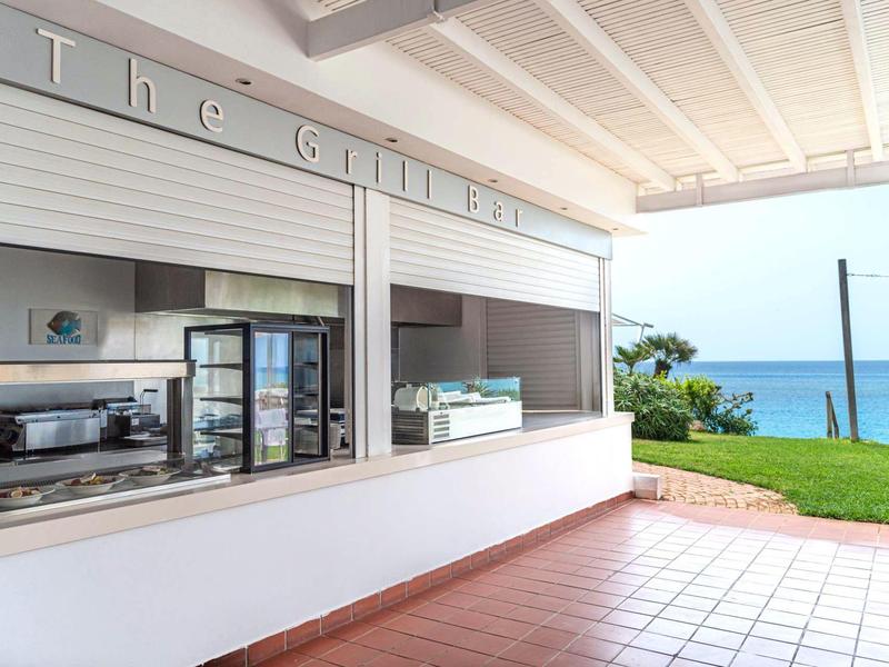 Offener Hotelbarbereich mit Blick auf einen Garten und das Meer im Hintergrund.