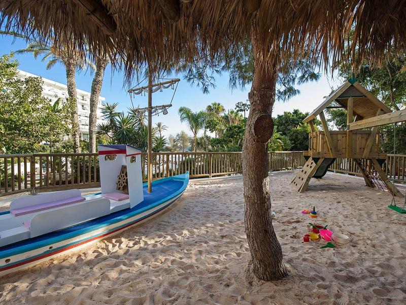 Spielplatz mit einem Boot, Rutsche und Schaukeln in einem sandigen Bereich unter Palmen.