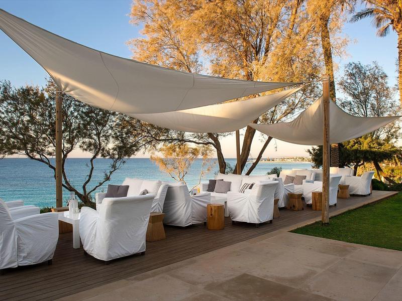 Außenbereich mit bequemen weißen Sesseln und Sonnensegeln neben Meer und Palmen.