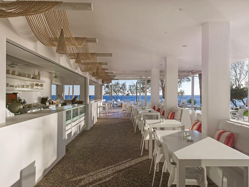 Helles, modernes Restaurant mit weißen Tischen und Stühlen, Blick auf den Pool und Palmen.