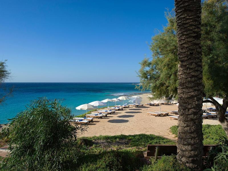Sandstrand mit blauen Sonnenschirmen und klarem türkisfarbenem Meer im Hintergrund