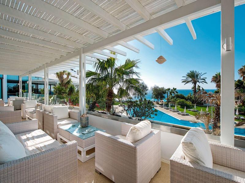 Luxuriöse Terrasse mit weißen Möbeln und Blick auf einen Pool und Palmen unter klarem Himmel.