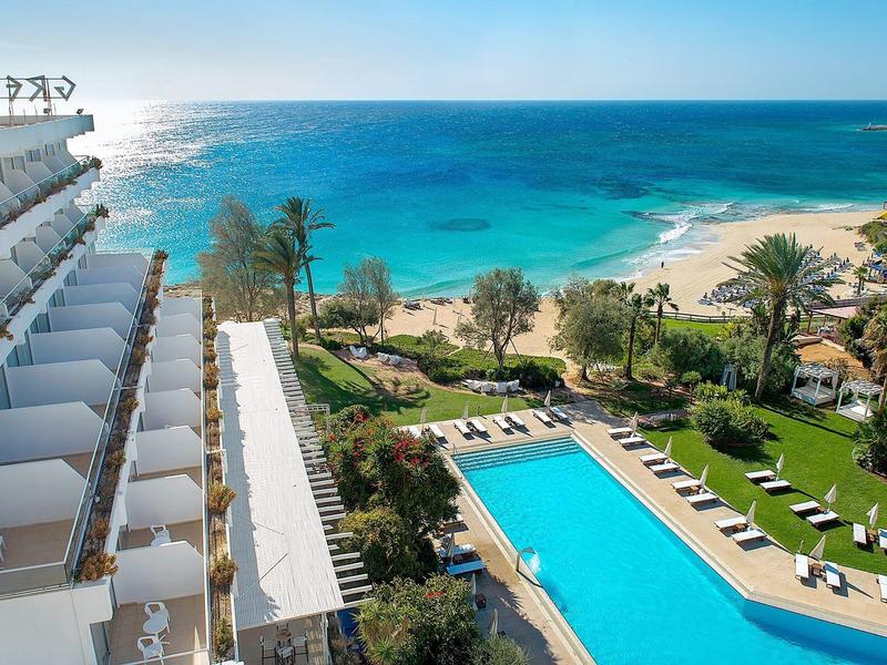 Hotel mit großem blauen Pool, grüner Garten, Palmen und Blick auf türkisfarbenen Ozean und Strand.