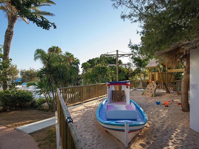 Ein kleines Spielboot im Sand neben einem weißen Gebäude und Palmen am Strand.