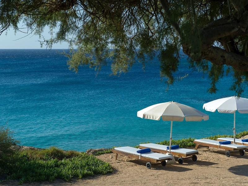 Strand mit Sonnenliegen und weißen Sonnenschirmen, dahinter klares blaues Meer und bewölkter Himmel.