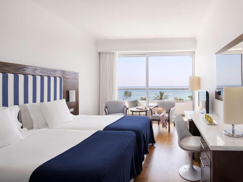 Hotelzimmer mit zwei Betten, blau-weißem Kopfteil und Meerblick durch großes Fenster.