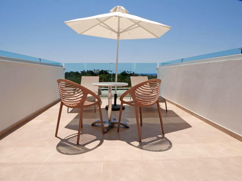 Terrasse mit zwei Stühlen, Tisch und weißem Sonnenschirm, Meer im Hintergrund bei klarem Himmel.