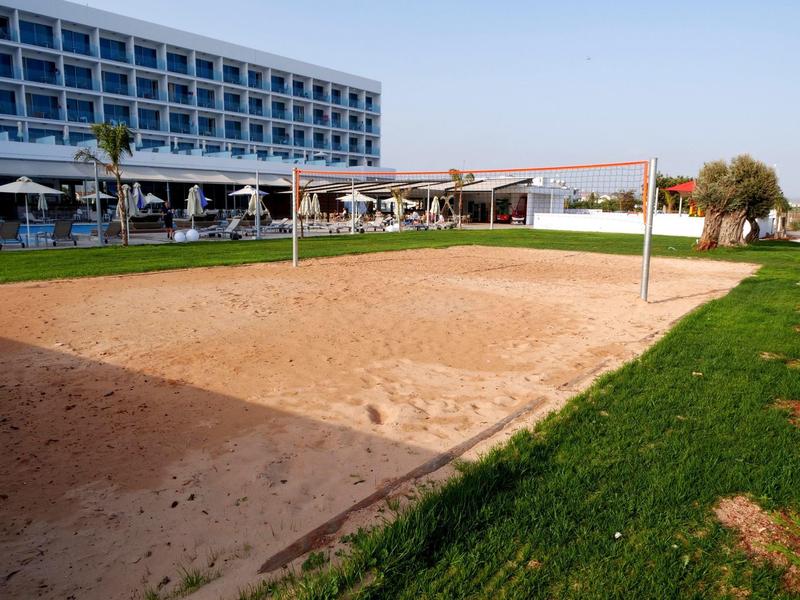 Sand-Volleyballfeld mit Netz, umgeben von grünem Rasen vor einem mehrstöckigen Hotelgebäude.