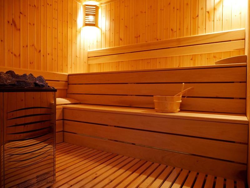Innenraum einer Sauna mit hölzernen Sitzbänken, Hocker und Beleuchtung an der Wand.
