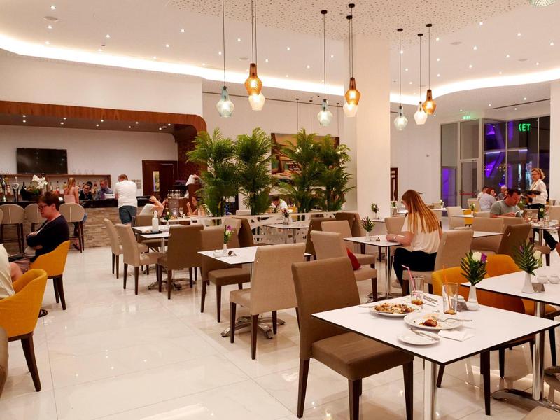 Moderne Restaurant mit weißen Böden, braunen Tischen, beigen Stühlen und warmem Deckenlicht.