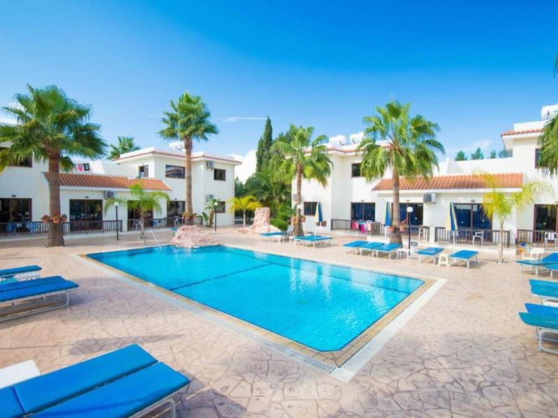 Schwimmbad mit blauem Wasser, umgeben von weißen Gebäuden und blauen Sonnenliegen unter Palmen.