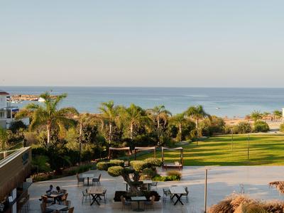 Blick auf eine Hotelanlage mit Garten, Sitzgelegenheiten und Meer im Hintergrund bei klarem Himmel.