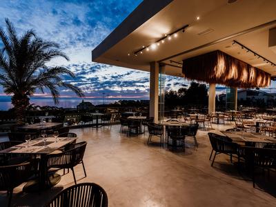 Offene Terrasse eines Hotels mit Tischen und Stühlen bei Sonnenuntergang am Meer.