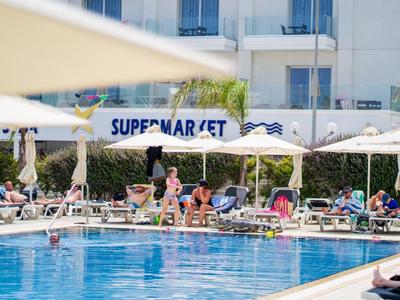 Hotelpool mit Sonnenliegen, Sonnenschirmen und Gästen neben einem Gebäude mit Supermarkt.