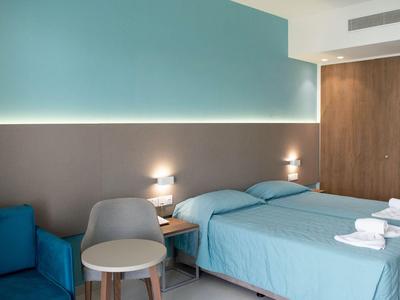 Modernes Hotelzimmer mit zwei Einzelbetten, blauem Sofa, Stuhl und Nachttisch unter indirekter Beleuchtung.