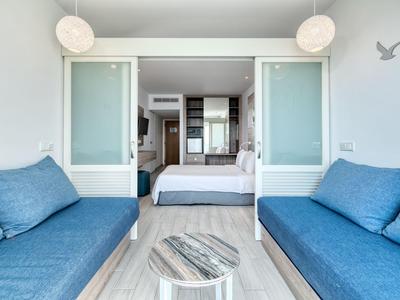 Modern eingerichtetes Hotelzimmer mit zwei blauen Sofas und einem Bett im Hintergrund.