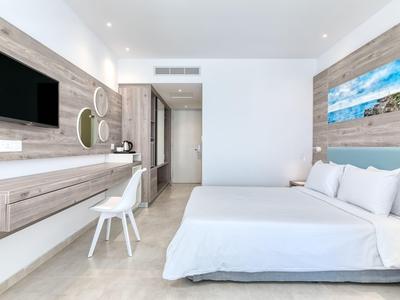 Camera d'hotel luminosa con letto matrimoniale, TV a parete e area di lavoro moderna con sedia.