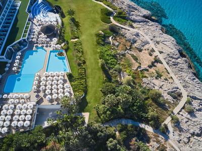 Vista aerea di un hotel con piscine, lettini, aree verdi e costa rocciosa vicino al mare.