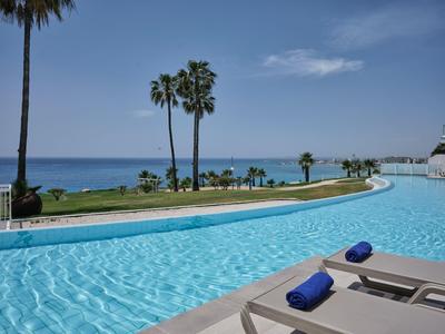 Una grande piscina curva con lettini e asciugamani, palme e mare sullo sfondo.