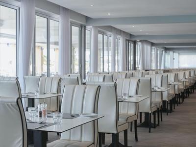 Sala da pranzo moderna con file di sedie imbottite bianche e tavoli accanto a grandi finestre.